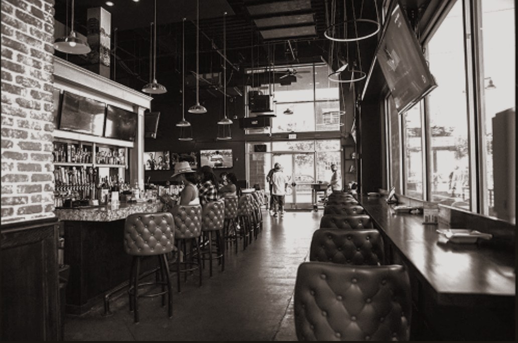 Long view down the bar at Tisdale's Cigar Lounge with tufted leather stools, a brick wall, and a counter along the front windows
